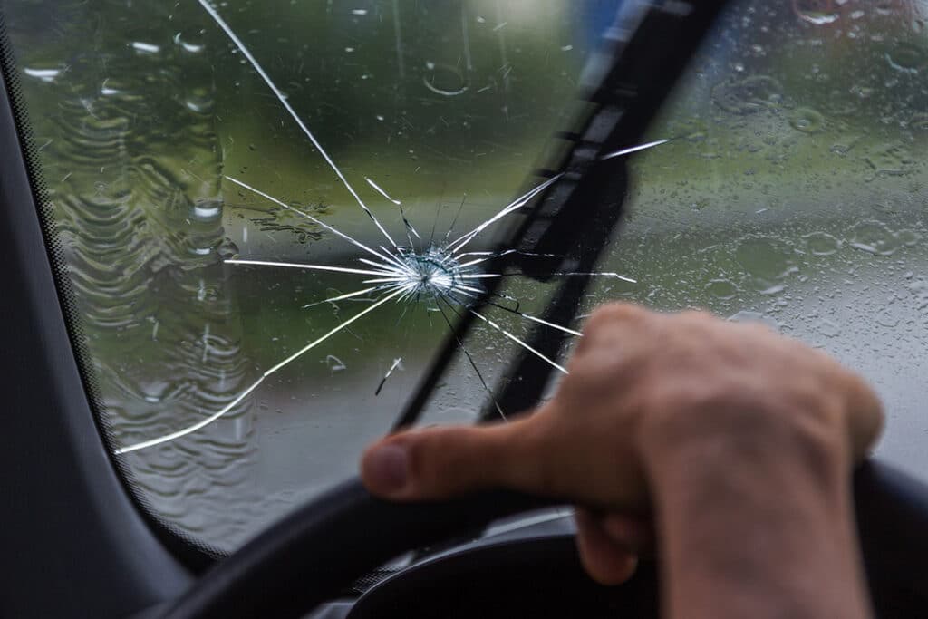 A close-up view of a cracked windshield with a visible star-shaped break while a hand grips the steering wheel. Rain is falling on the glass, creating streaks and droplets.
